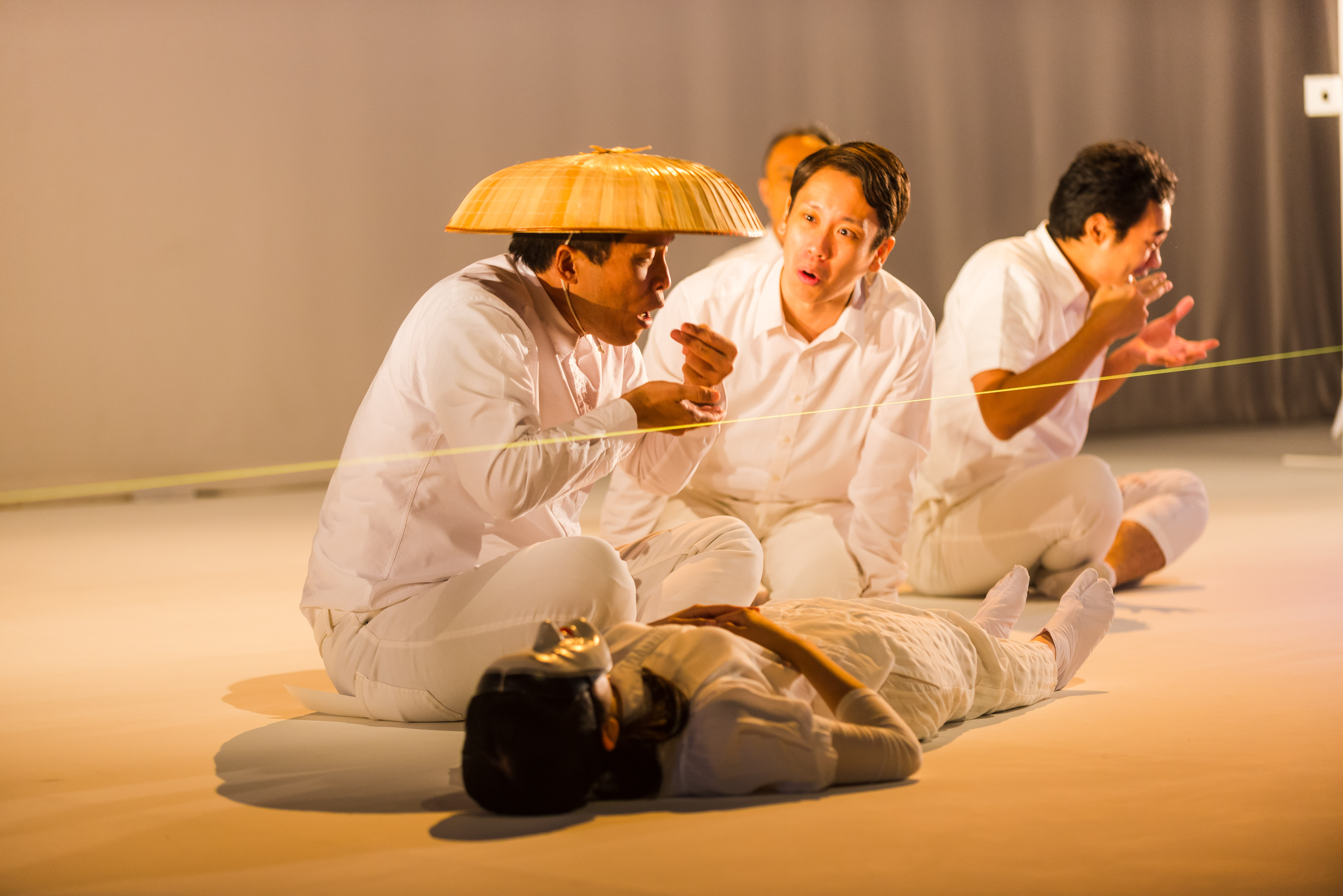 A production still from SEED (2015). A body lies on the stage floor. Beside it sits 3 male performers, dressed in white shirt and pants. 2 of them hold up their hands to their mouths as if miming eating from a bowl, while the third who is in the middle looks to them questioningly.