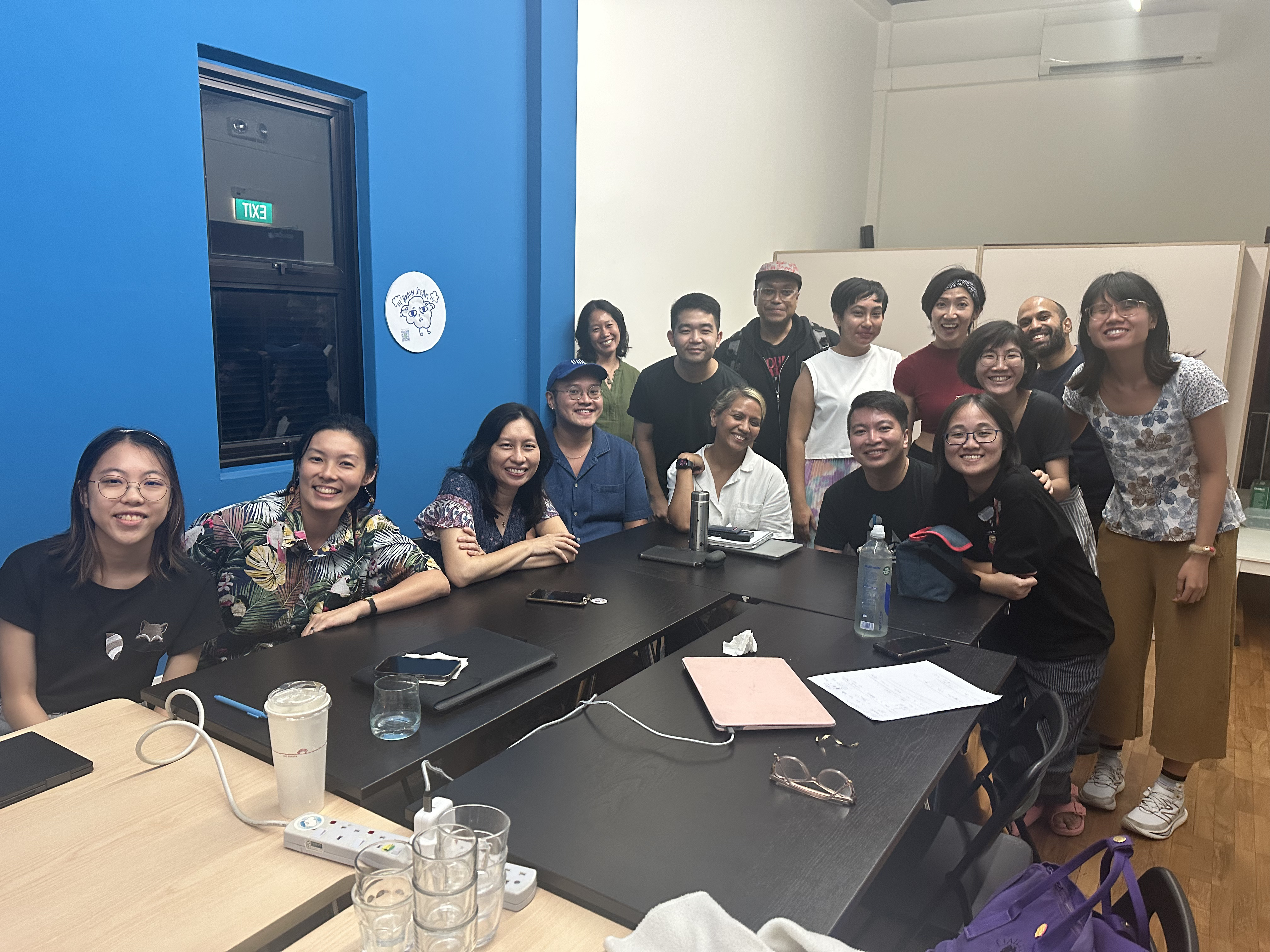 A group photo of 15 people seated and standing around rectangular tables, smiling.