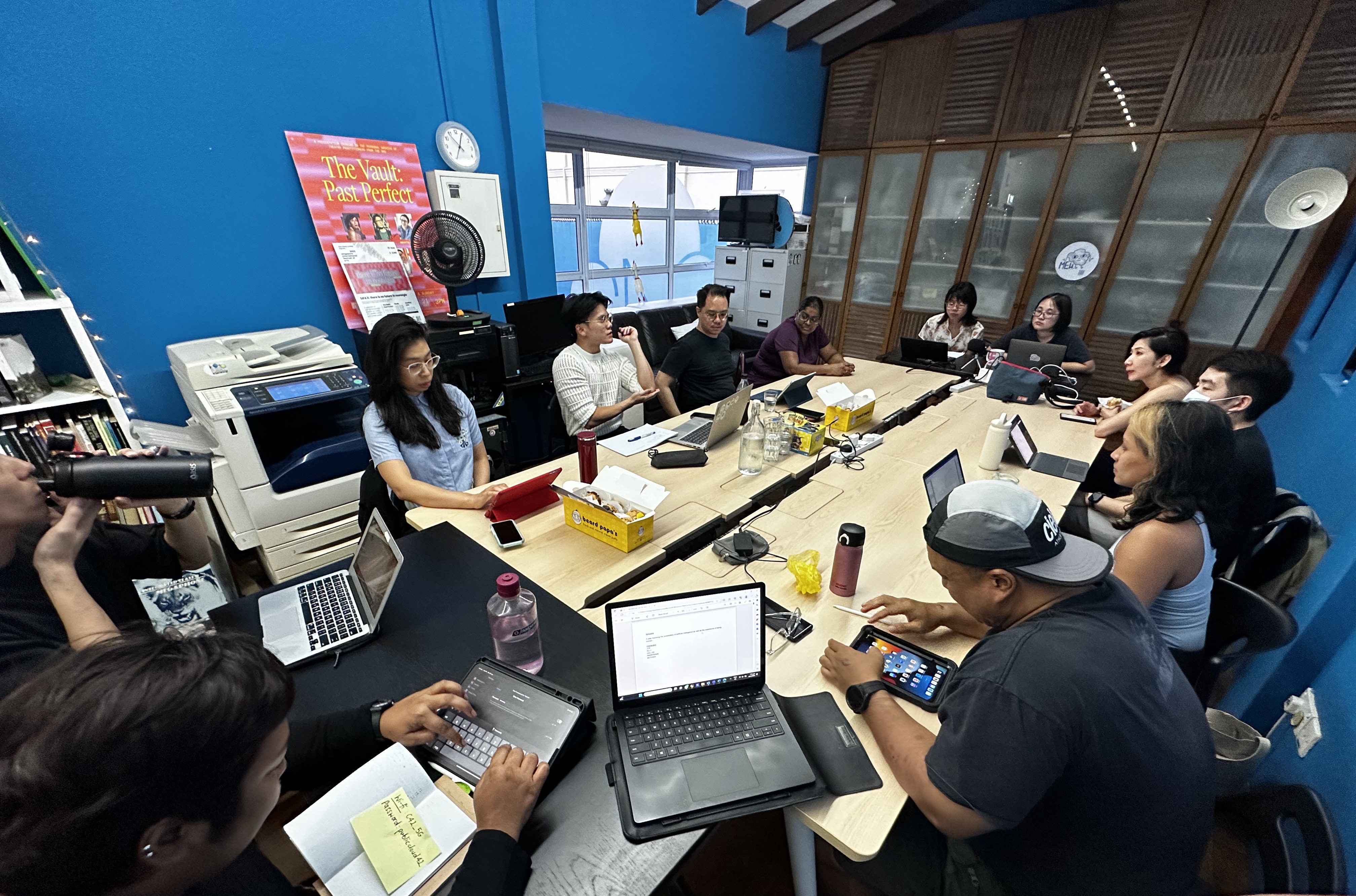 Twelve people seated around tables in a rectangular formation, with scripts and laptops in front of them.