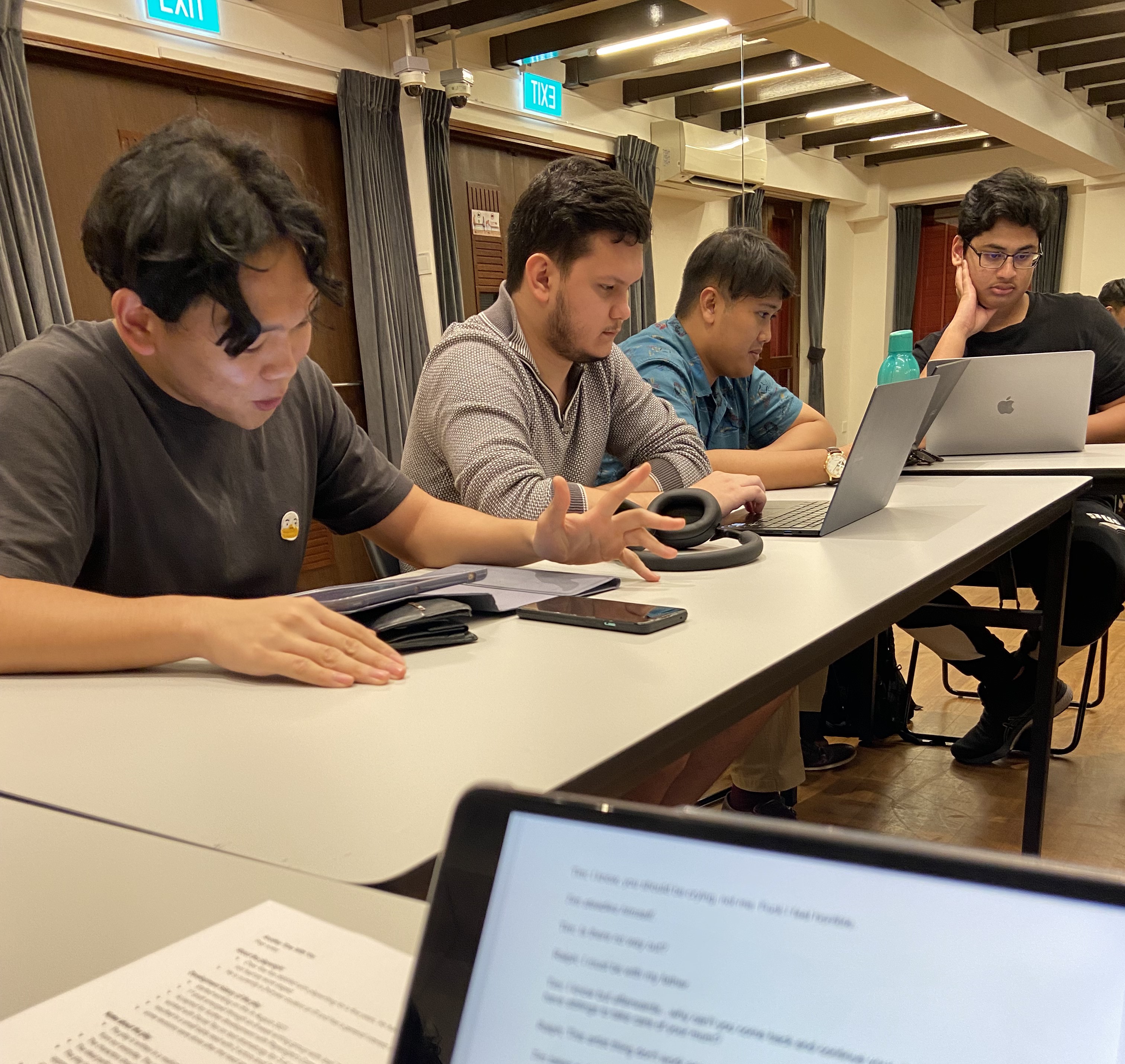 A photograph with Marwyn, Christopher, Rifqi and Rohan seated in front of a long grey table, with a digital and physical copy of a script in the foreground.