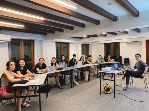 A group of people seated along a row of tables in the 42 Waterloo Street Rehearsal Studio. In front of them is another person seated behind a single table, on which is a laptop and table. Everyone is smiling at the camera.