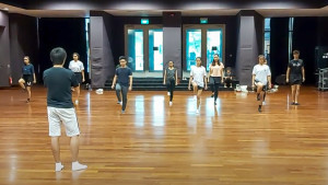 A group of people standing in a large, bright room with wooden flooring, raising their right legs. Standing in front of them, with his back to the camera, is an instructor with his arms folded.